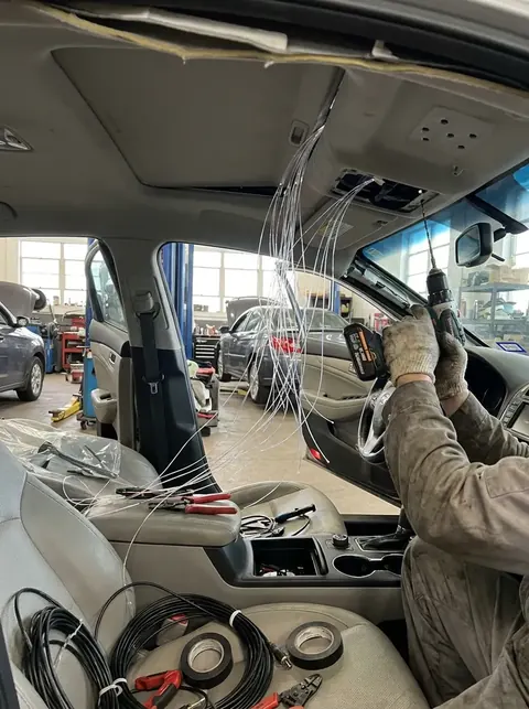 Render of a car in a workshop with the roof lining pulled down, loose fibre optic strands hanging and a mechanic holding a drill.
